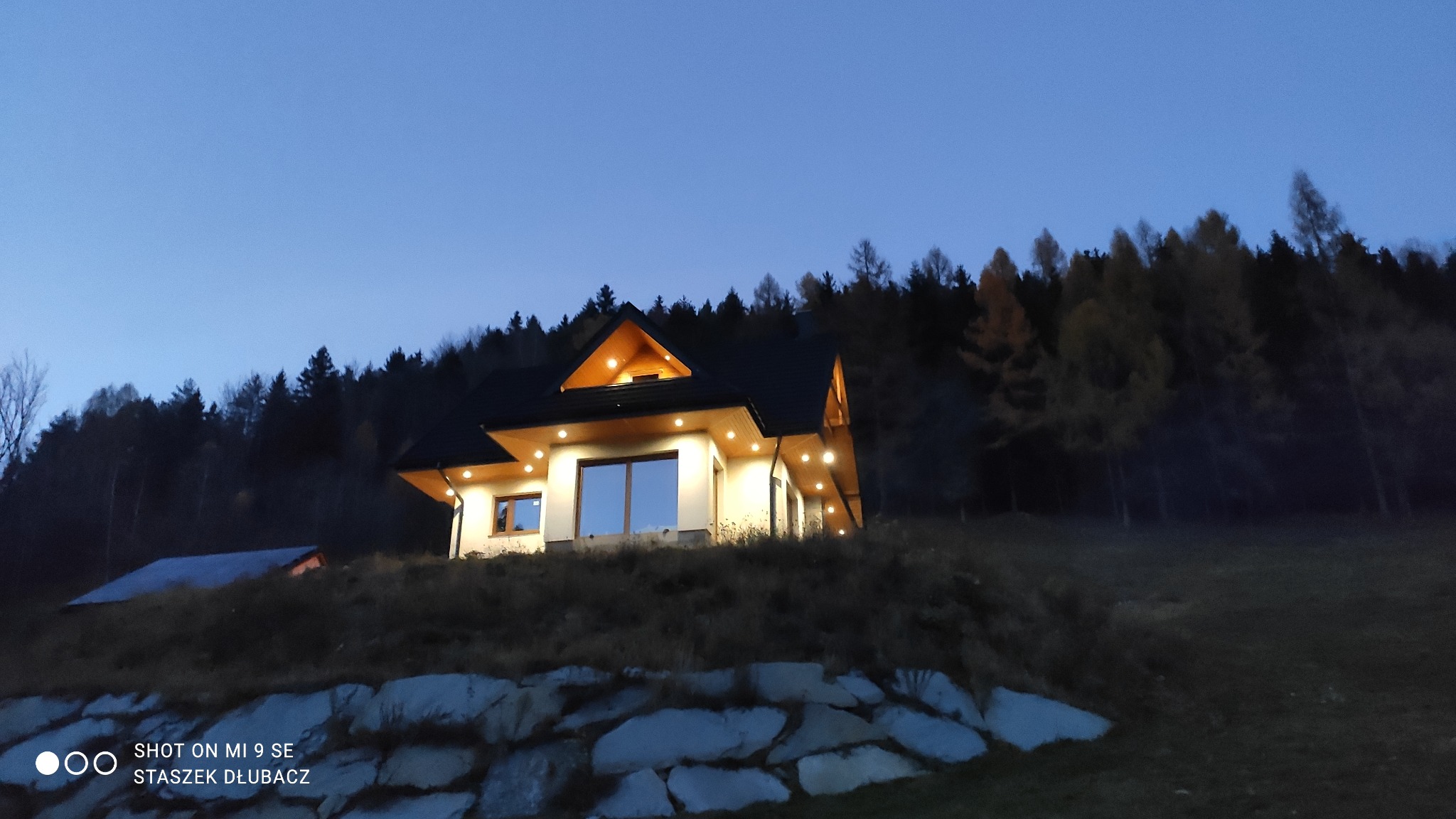 Oświetlony dom na wzgórzu o zmierzchu, z widocznym kamiennym murem oporowym i lasem w tle.
