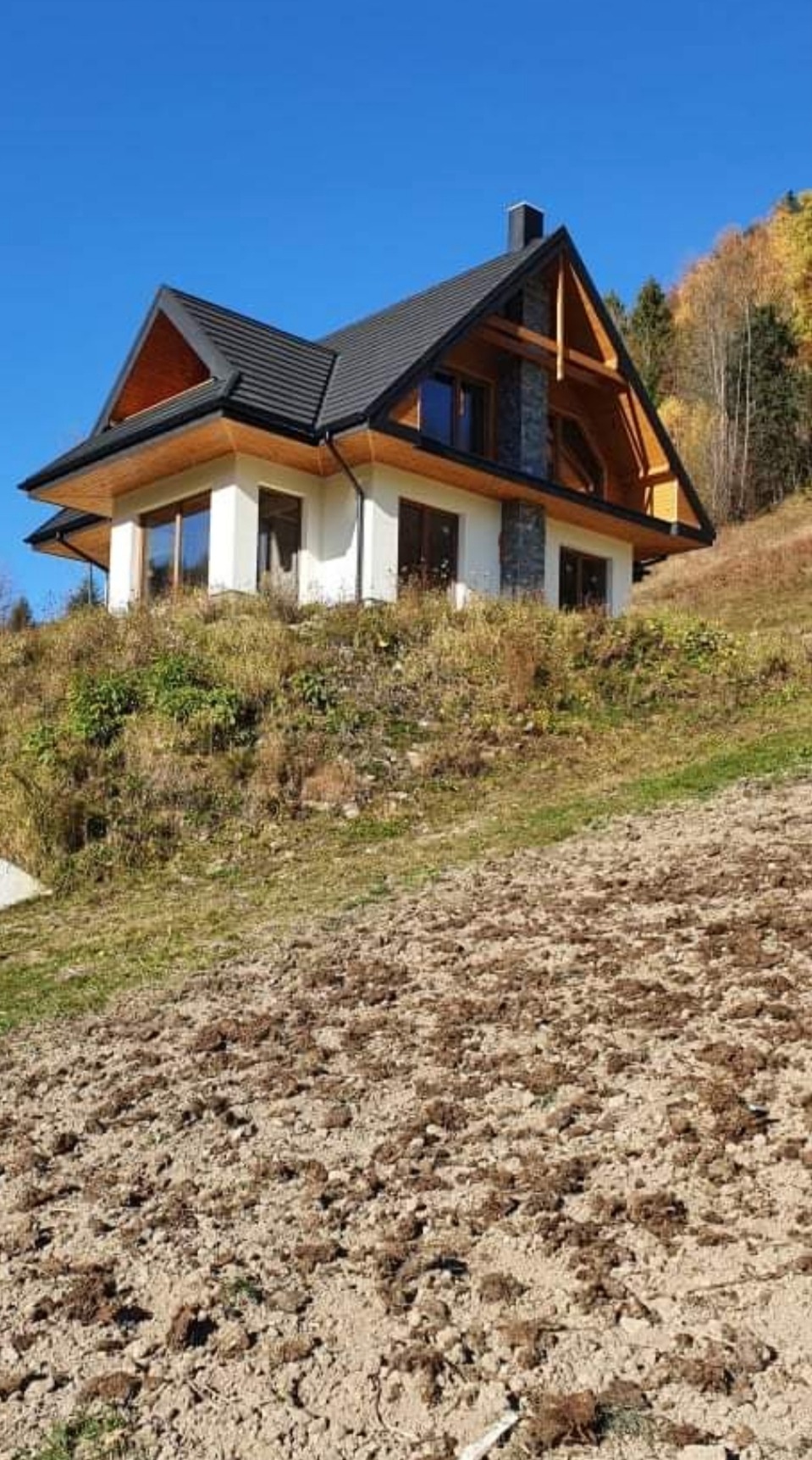 Nowoczesny dom z jasną elewacją i ciemną dachówką, położony na zboczu wzgórza z przygotowanym terenem pod ogród.