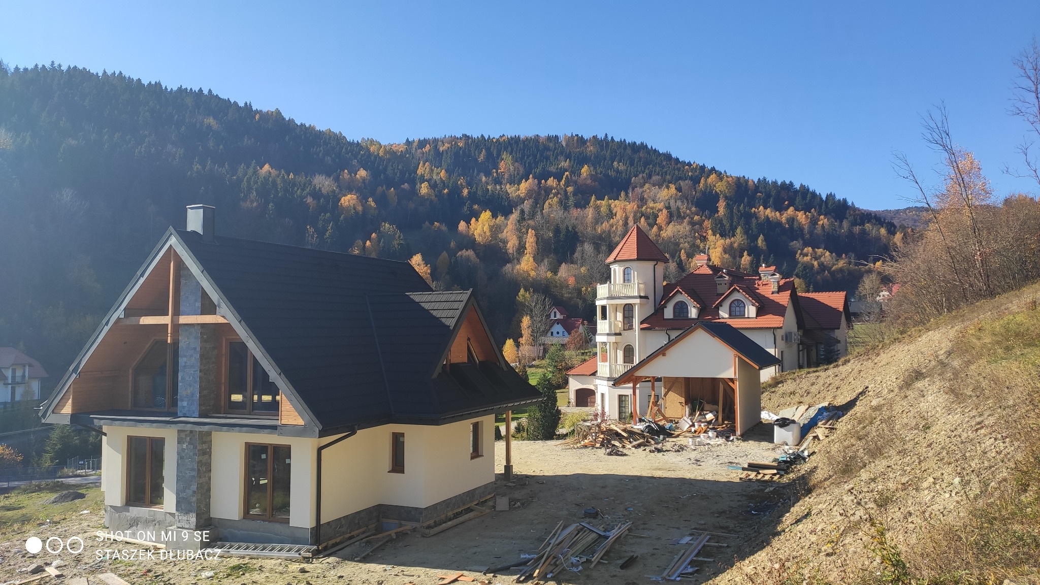 Niedokończony dom z drewnianą konstrukcją w stylu góralskim na tle jesiennego lasu i wieży mieszkalnej z balkonami.