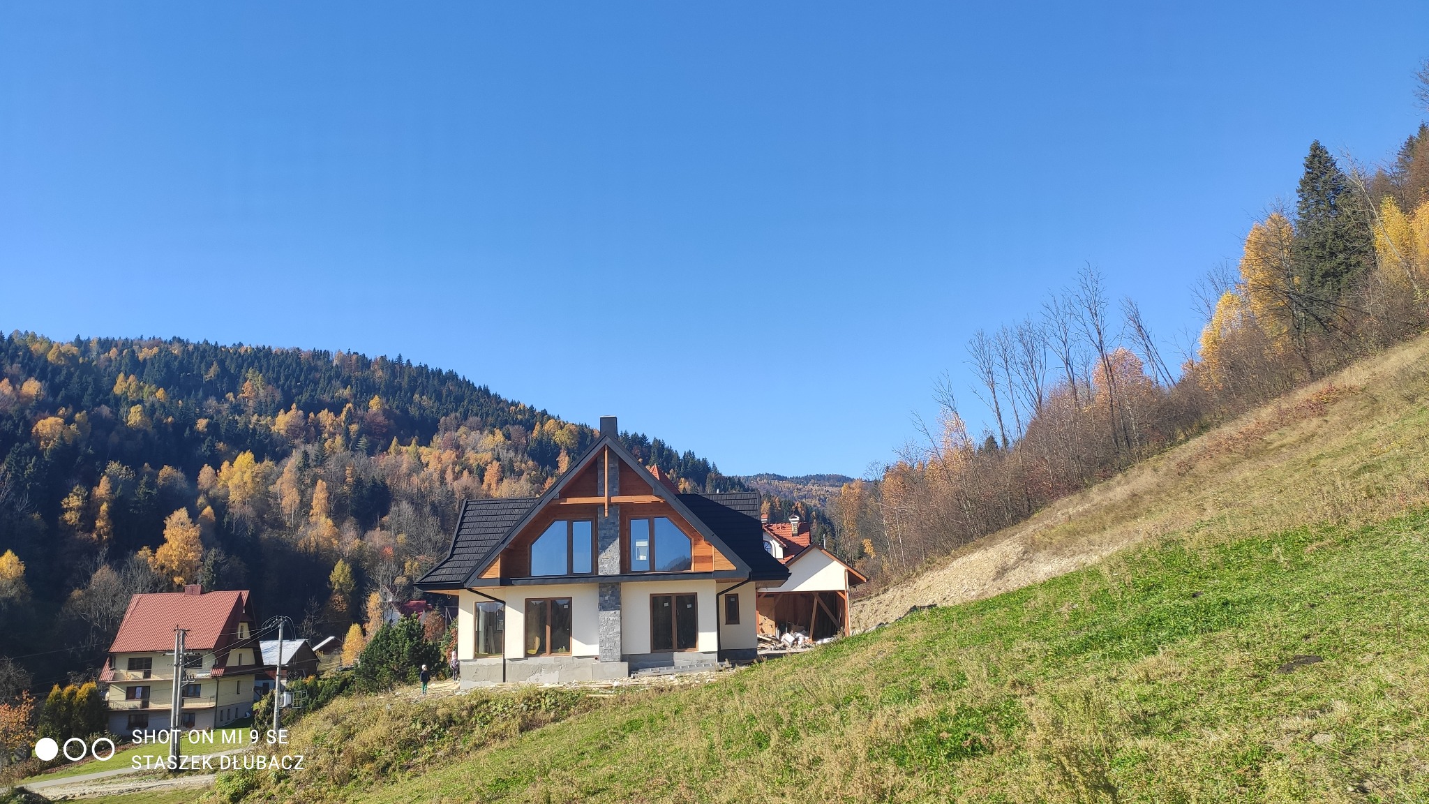 Nowoczesny dom z dużymi oknami, czarnym dachem i drewnianymi elementami elewacji, położony na wzgórzu z widokiem na las w jesiennych barwach.