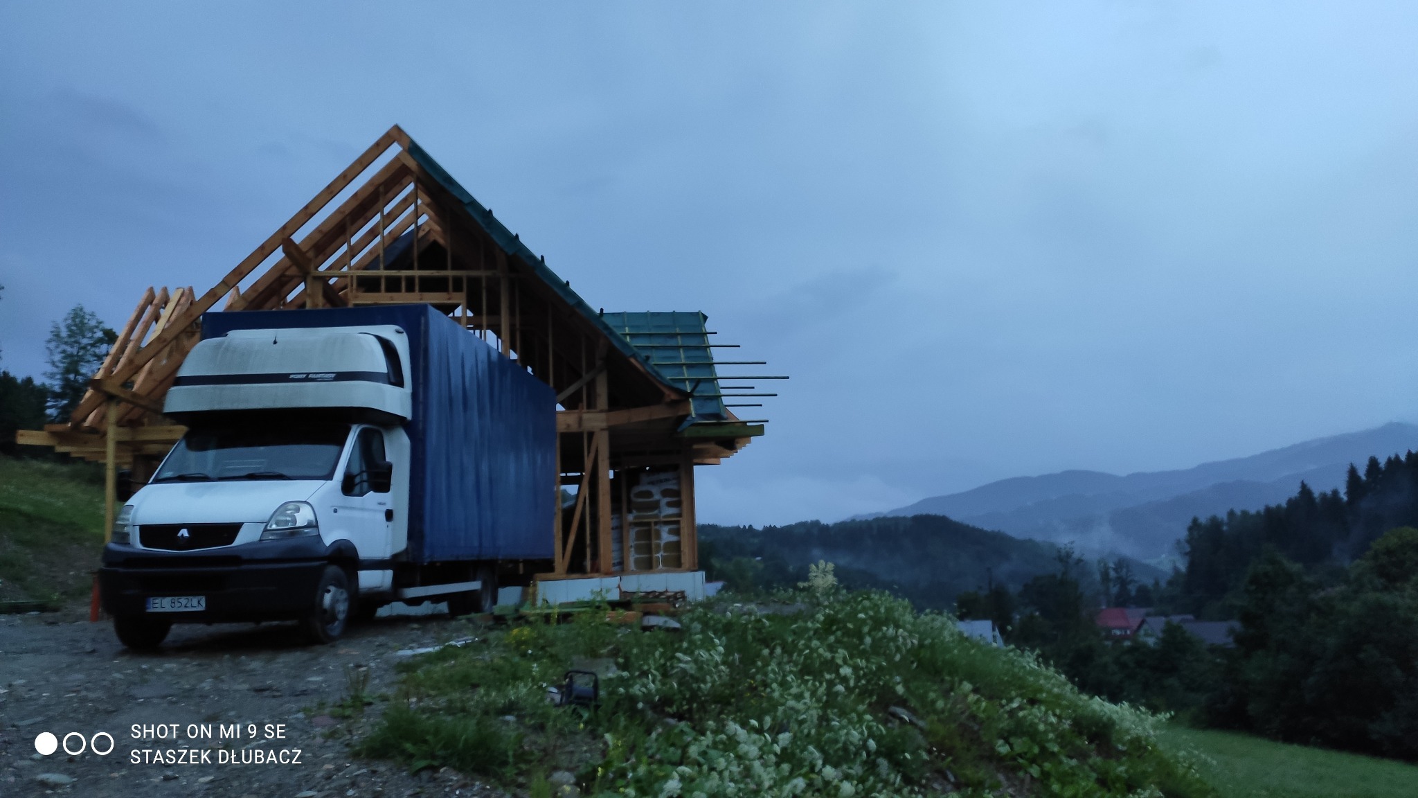 Wczesny etap budowy domu z drewnianą konstrukcją szkieletową, z dostawczym autem na pierwszym planie i górskim krajobrazem w tle.