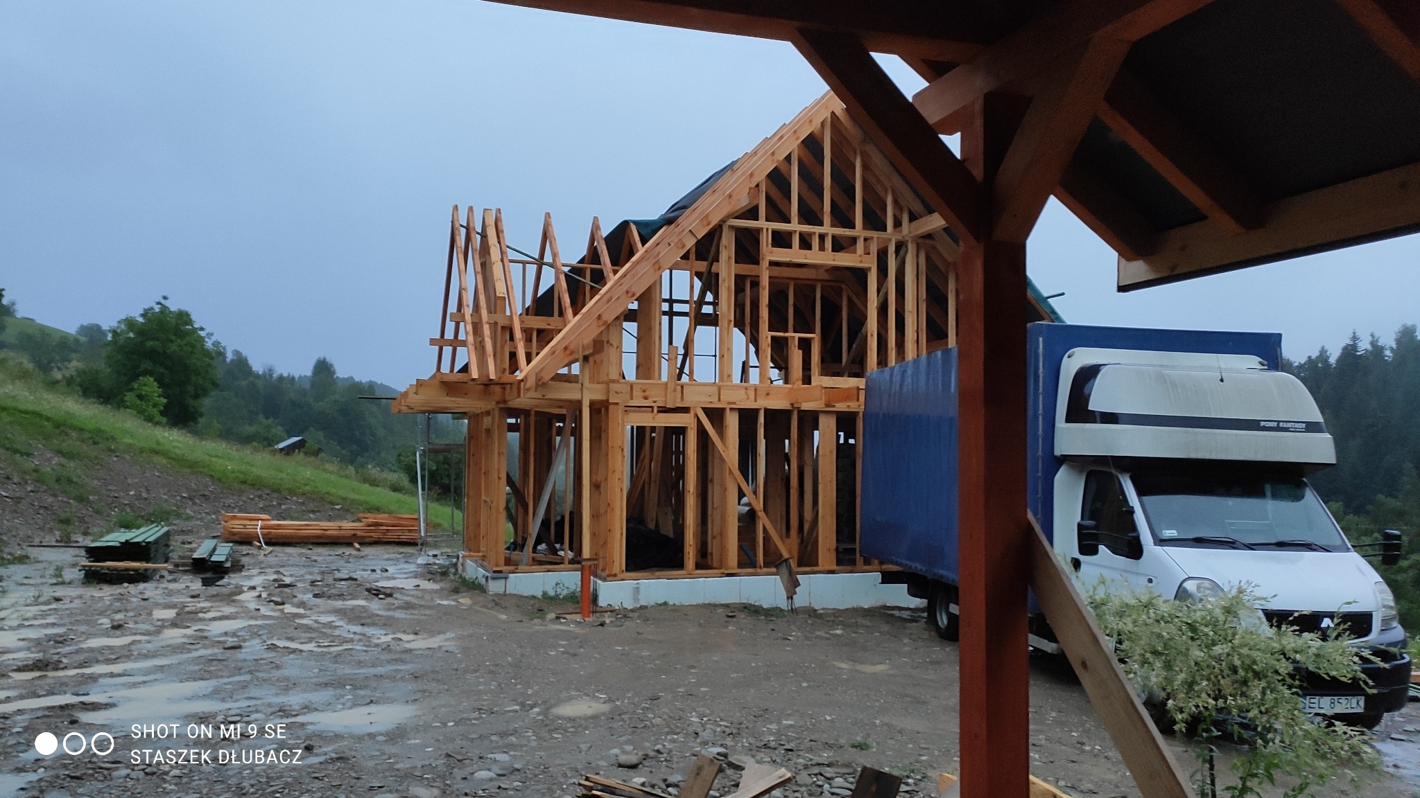 Surowa konstrukcja domu w trakcie budowy, widoczna drewniana rama i elementy dachu, na zewnątrz zaparkowany biały samochód dostawczy z niebieską plandeką, teren budowy błotnisty po deszczu, w tle...