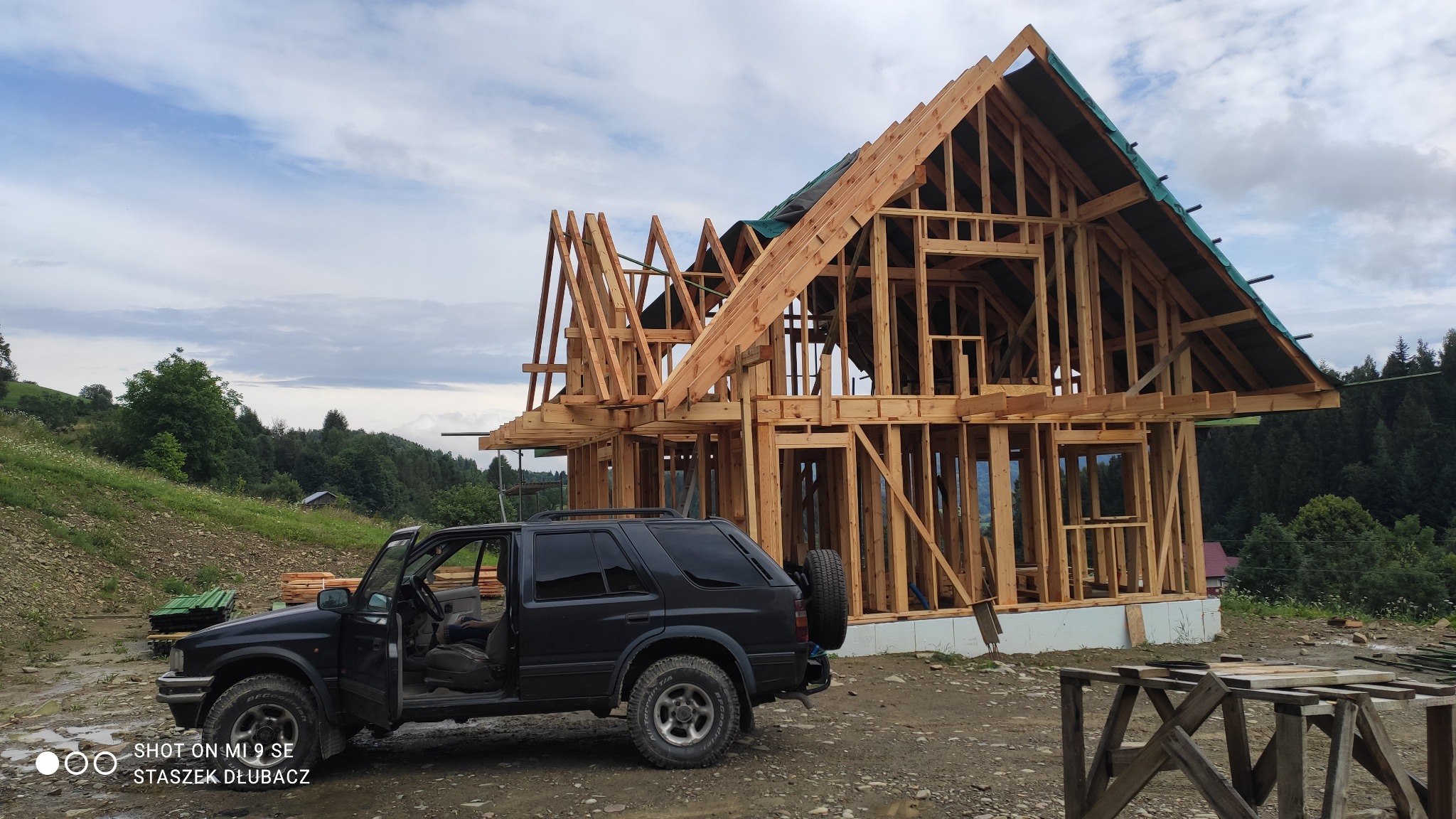 Konstrukcja drewniana domu w trakcie budowy, widoczny szkielet budynku z otwartymi drzwiami czarnego samochodu terenowego na pierwszym planie, górzysty krajobraz w tle.
