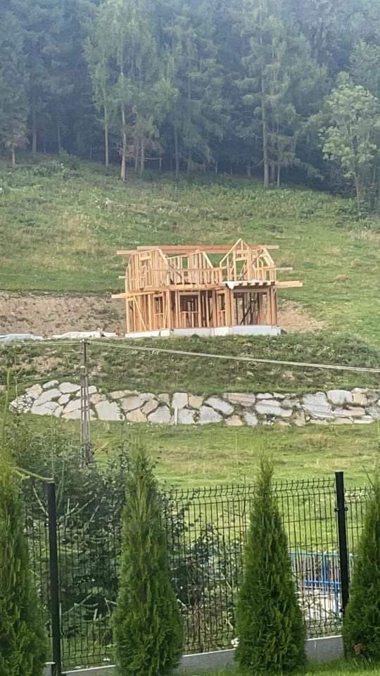 Konstrukcja domu szkieletowego w trakcie budowy na wzgórzu, widok zza ogrodzenia i drzew iglastych.