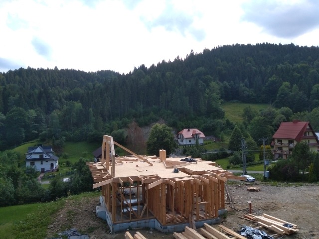 Wznoszenie konstrukcji drewnianej domu w górskim otoczeniu, widoczne fundamenty i elementy szkieletu, w tle zalesione wzgórza i zabudowa mieszkalna.
