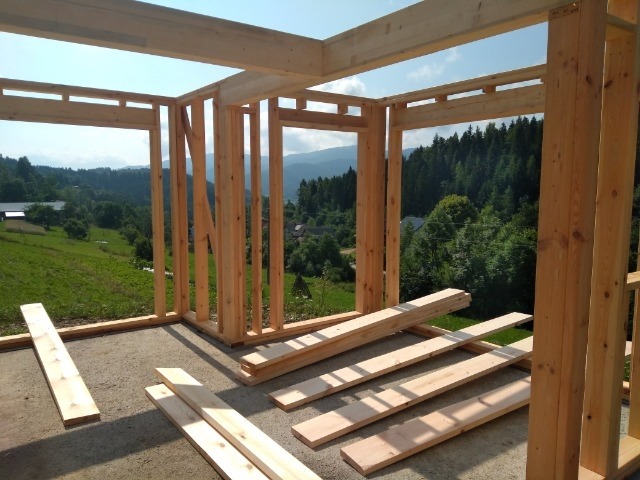Wznoszenie konstrukcji drewnianej z widokiem na zielone wzgórza i lasy w tle, elementy konstrukcyjne ułożone na podłodze.