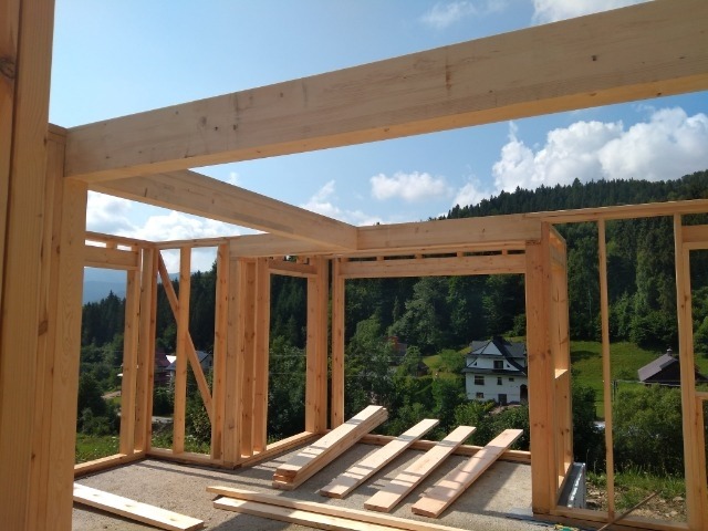 Wnętrze surowego stanu budynku z drewnianą konstrukcją szkieletową, widok na zalesione wzgórza i błękitne niebo.