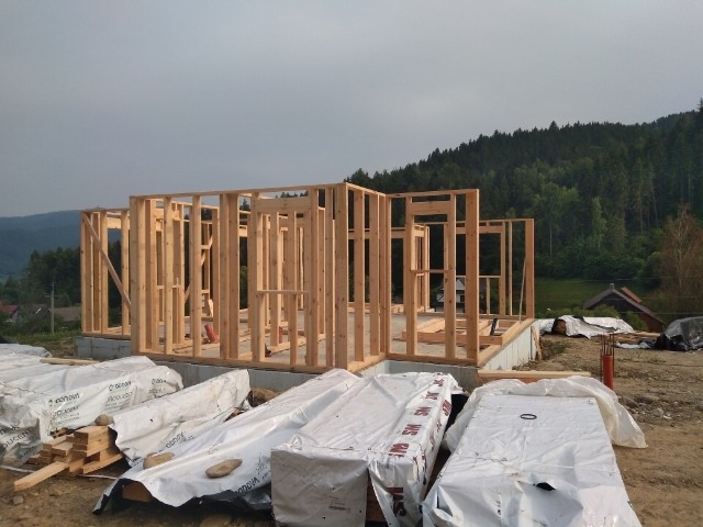 Konstrukcja szkieletowa domu w trakcie budowy na fundamencie, z widocznymi belkami i przygotowanymi materiałami budowlanymi w folii, na tle zalesionego wzgórza.