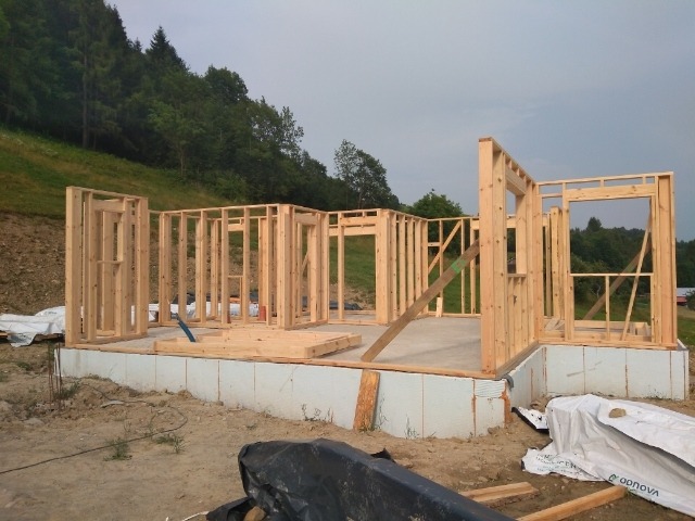 Konstrukcja domu szkieletowego w trakcie budowy, widoczne drewniane ramy ścian na fundamencie z izolacją, w tle zalesione wzgórze.