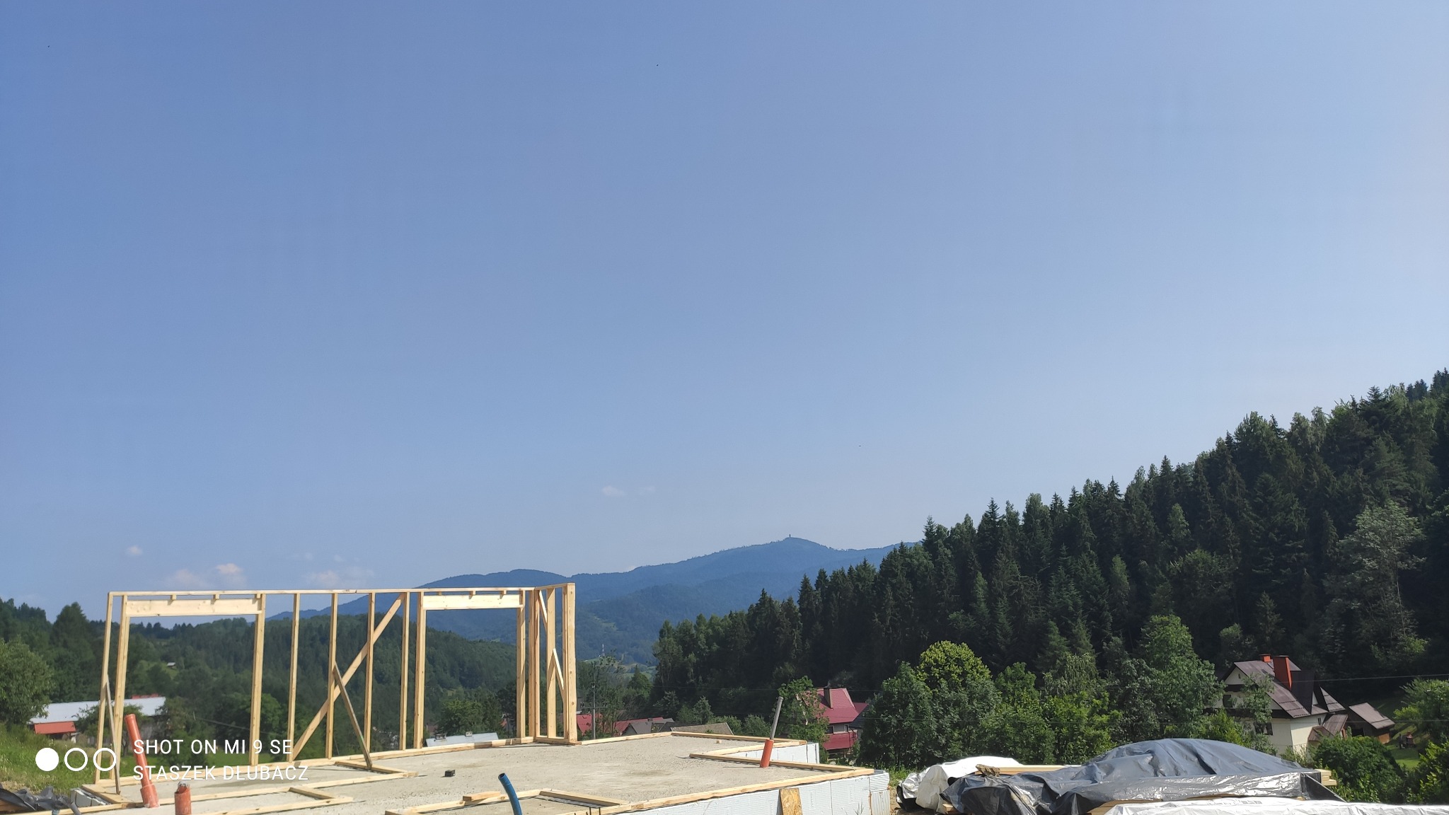 Wznoszenie drewnianej konstrukcji szkieletowej domu na betonowej płycie fundamentowej, w tle zalesione wzgórza pod błękitnym niebem.