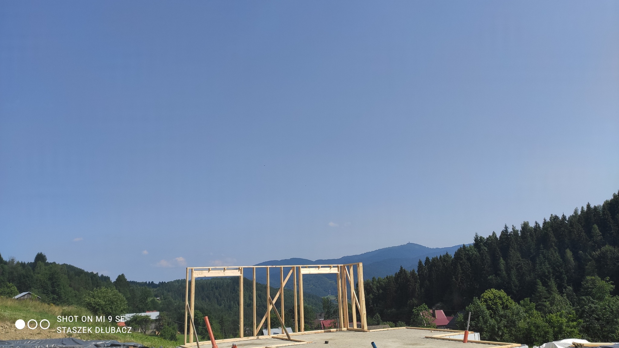 Wznoszenie drewnianej konstrukcji domu w malowniczym, górskim otoczeniu, widok na szczyt góry w tle.