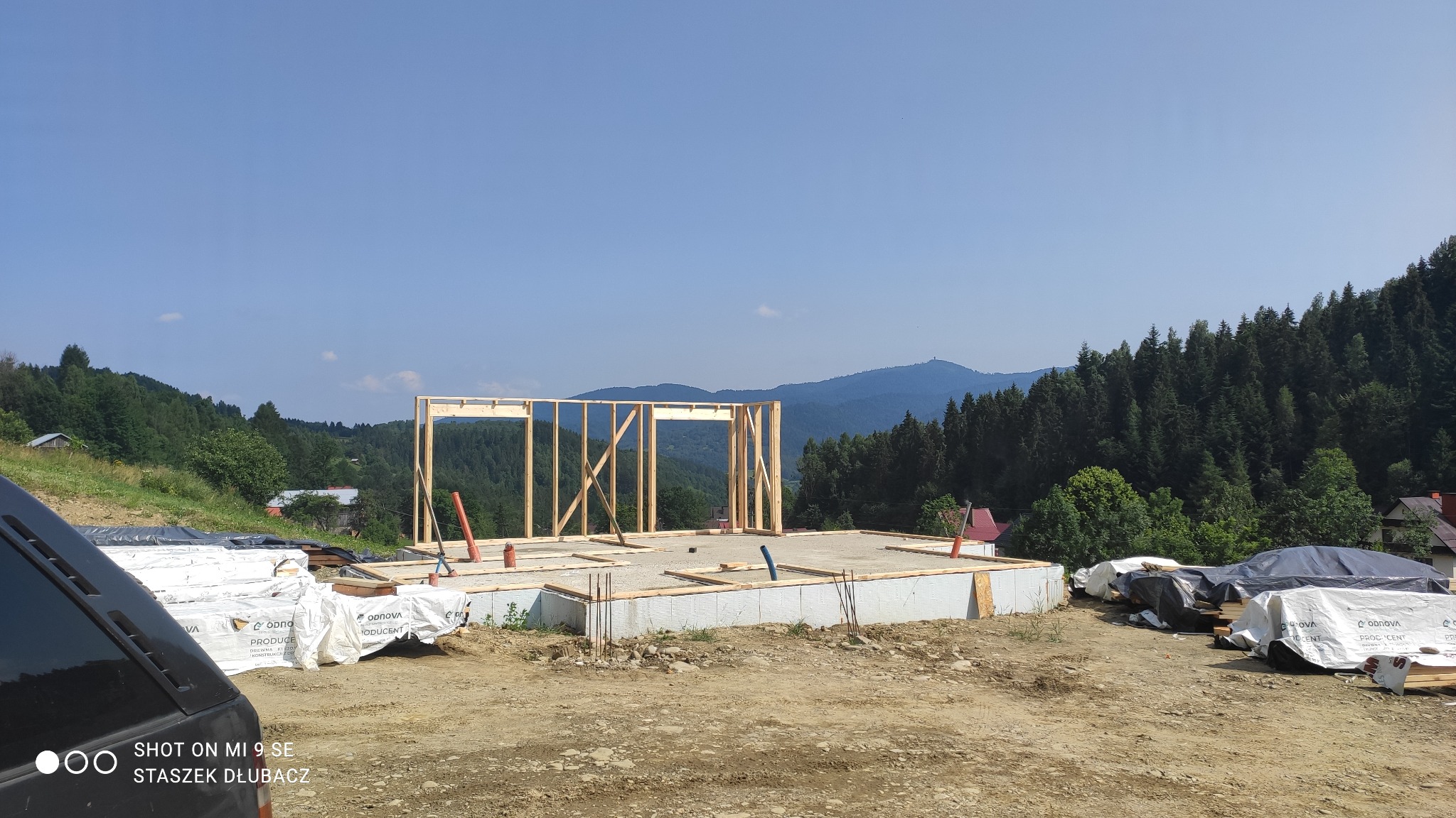 Wznoszenie konstrukcji ścian parteru na betonowej płycie fundamentowej w otoczeniu górskiego krajobrazu, widoczne materiały budowlane zabezpieczone folią.