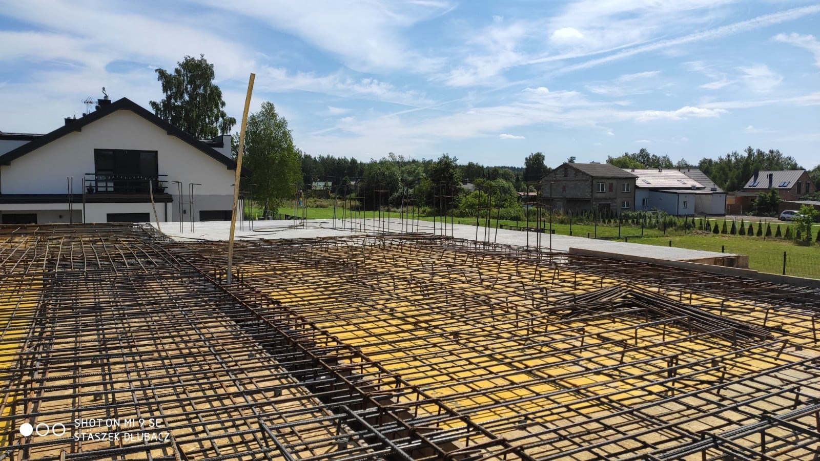 Zbrojenie płyty fundamentowej domu szkieletowego z widocznym deskowaniem i wystającymi prętami, w tle zabudowa jednorodzinna i błękitne niebo.