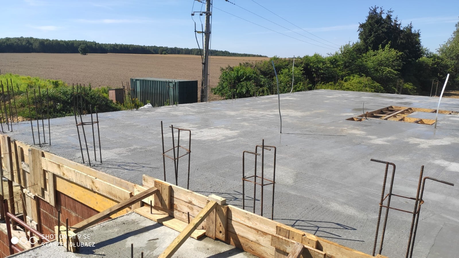 Wylany fundament z wystającymi prętami zbrojeniowymi i drewnianym szalunkiem, w tle pole i zieleń.