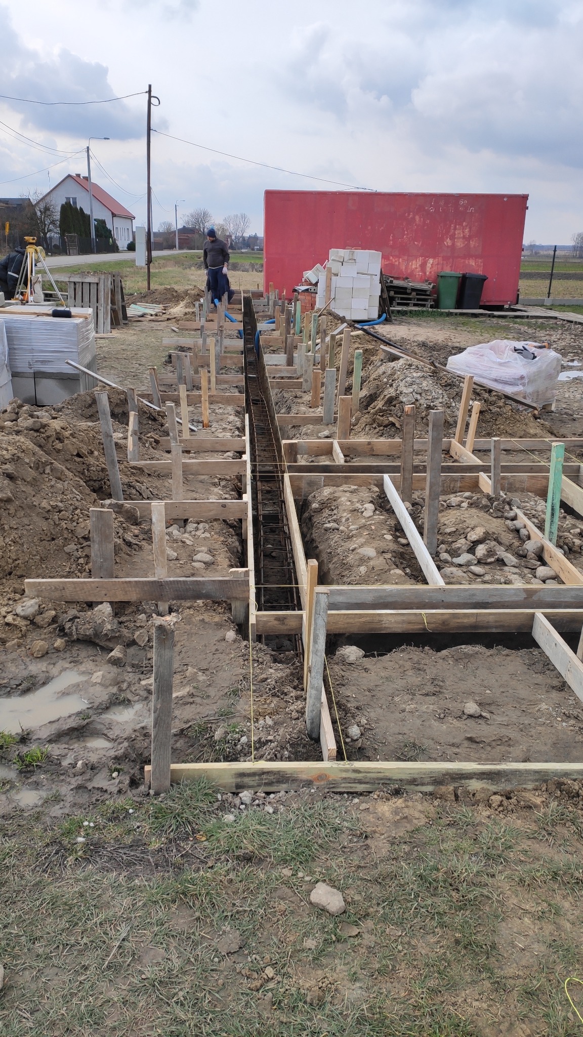 Przygotowanie fundamentów pod budowę: szalunki drewniane, zbrojenie stalowe, widoczny pracownik w oddali, teren budowy z błotem i elementami konstrukcyjnymi.