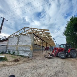 Radosław Wojtachnio - Budowa wiaty z drewnianą konstrukcją dachu, widoczny czerwony traktor z przodu i słup wysokiego napięcia po lewej stronie.