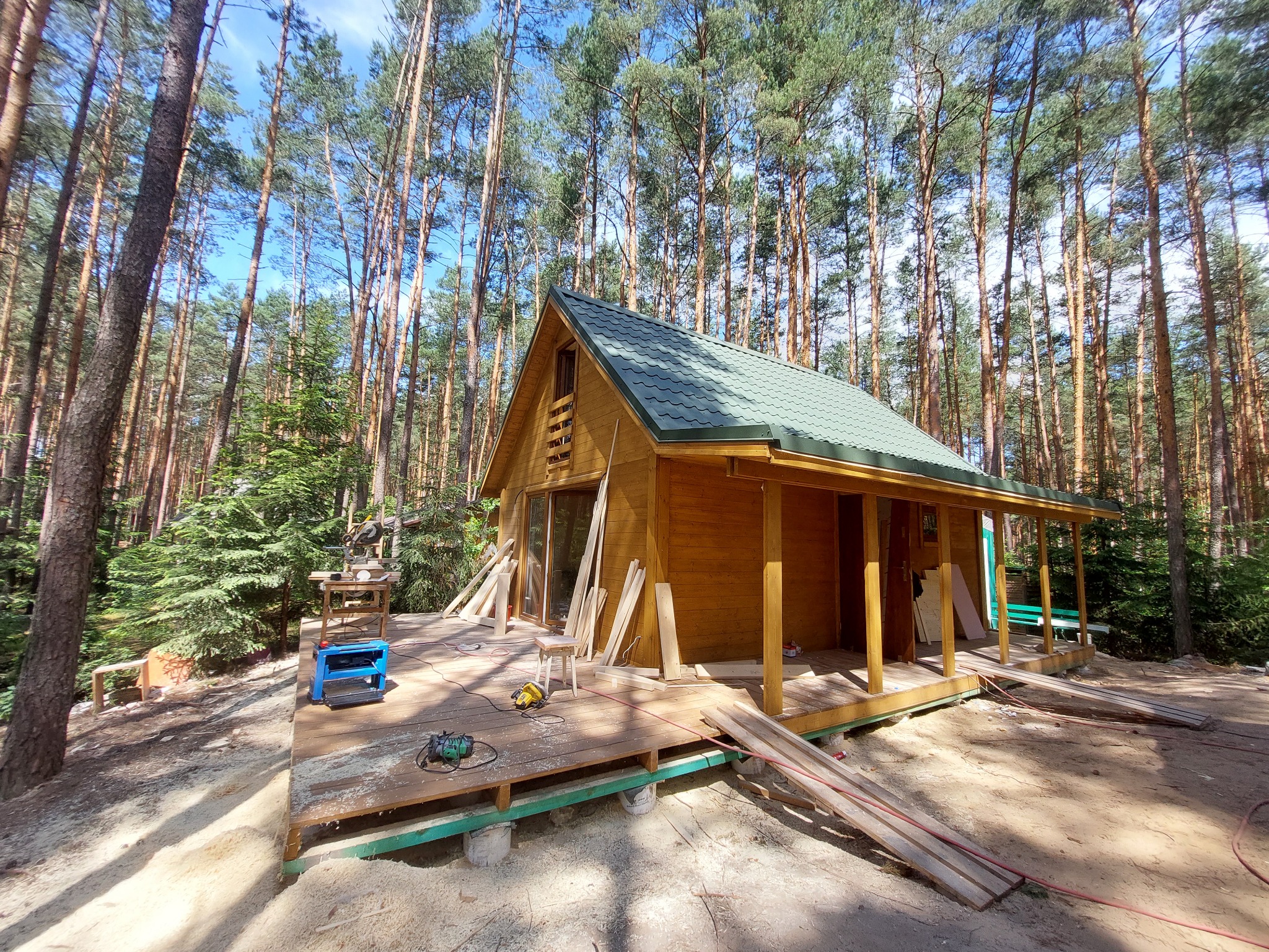 Drewniany domek letniskowy z zielonym dachem w trakcie budowy na drewnianym podeście, otoczony sosnowym lasem