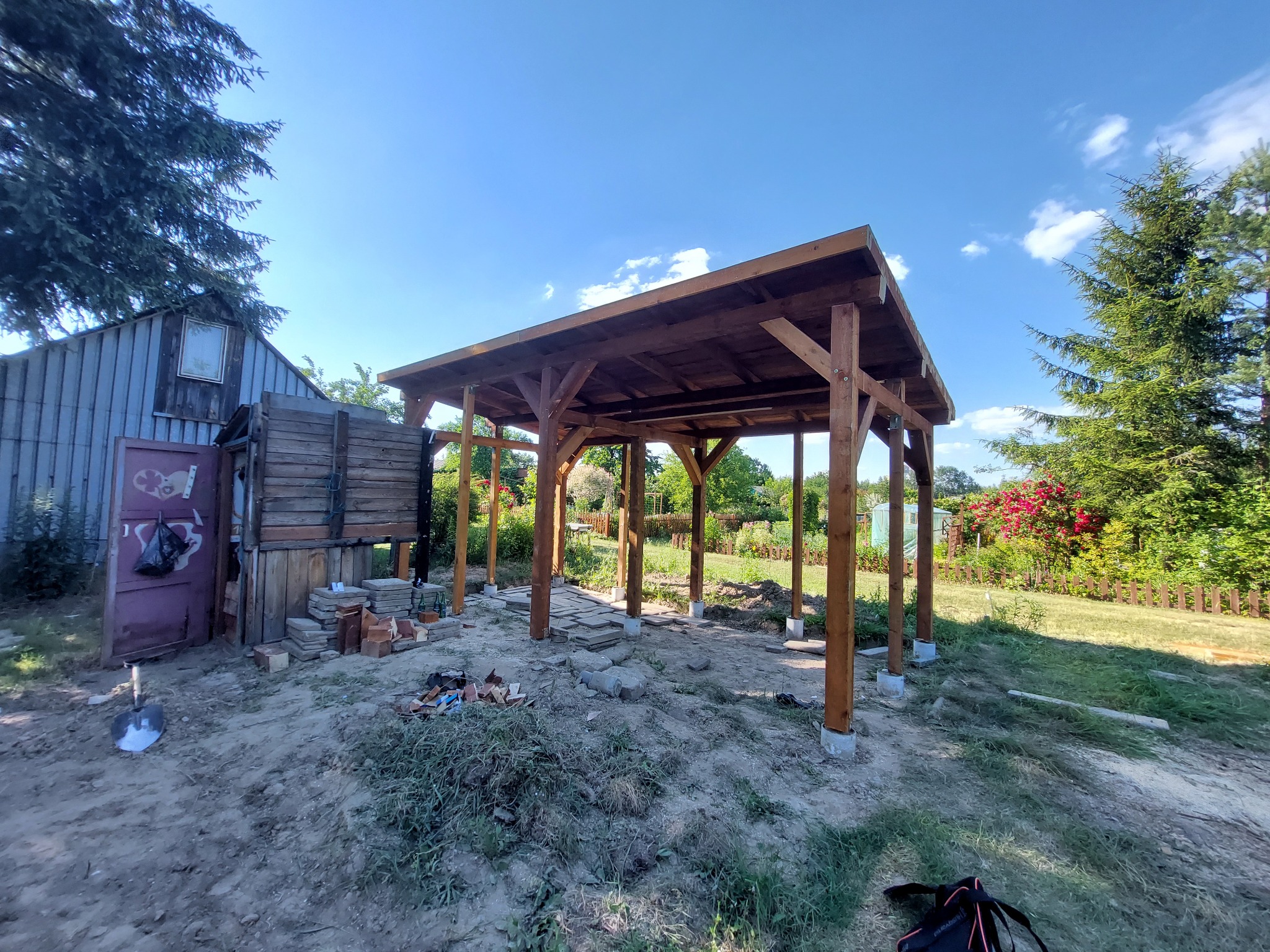 Drewniana konstrukcja zadaszenia w trakcie budowy, widoczne słupy, krokwie i deski dachowe, w tle ogród i budynek gospodarczy, na ziemi porozrzucane materiały budowlane.
