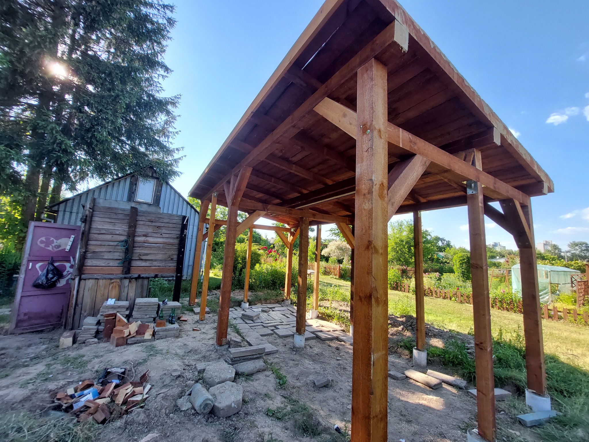 Konstrukcja drewnianej altany ogrodowej w trakcie budowy, widoczne słupy i krokwie, częściowo ułożona podłoga z płyt chodnikowych, w tle budynek gospodarczy.