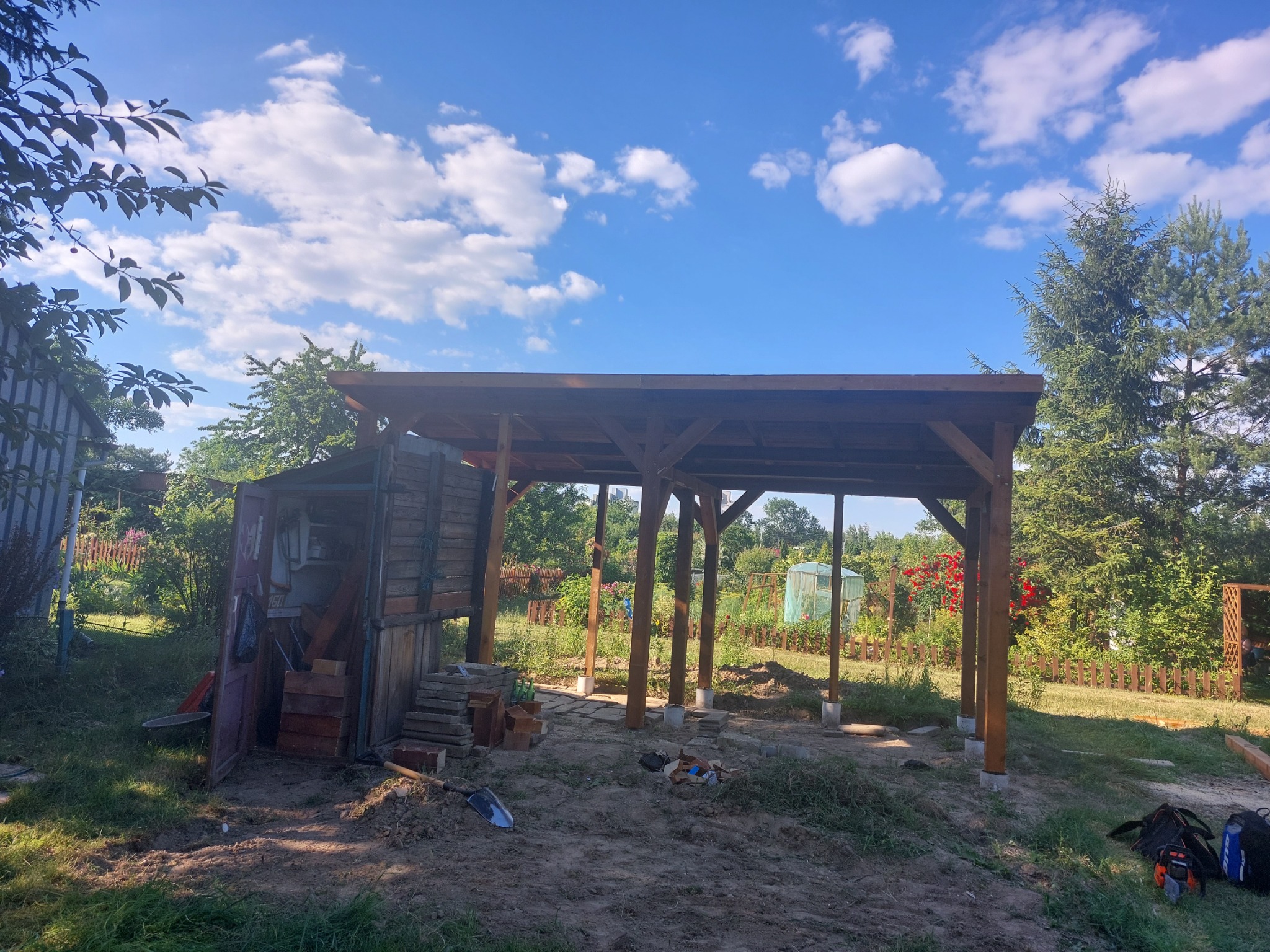 Drewniana konstrukcja zadaszenia w trakcie budowy, widoczne słupy i belka stropowa, narzędzia budowlane leżące na ziemi, tło z zielenią i błękitnym niebem.