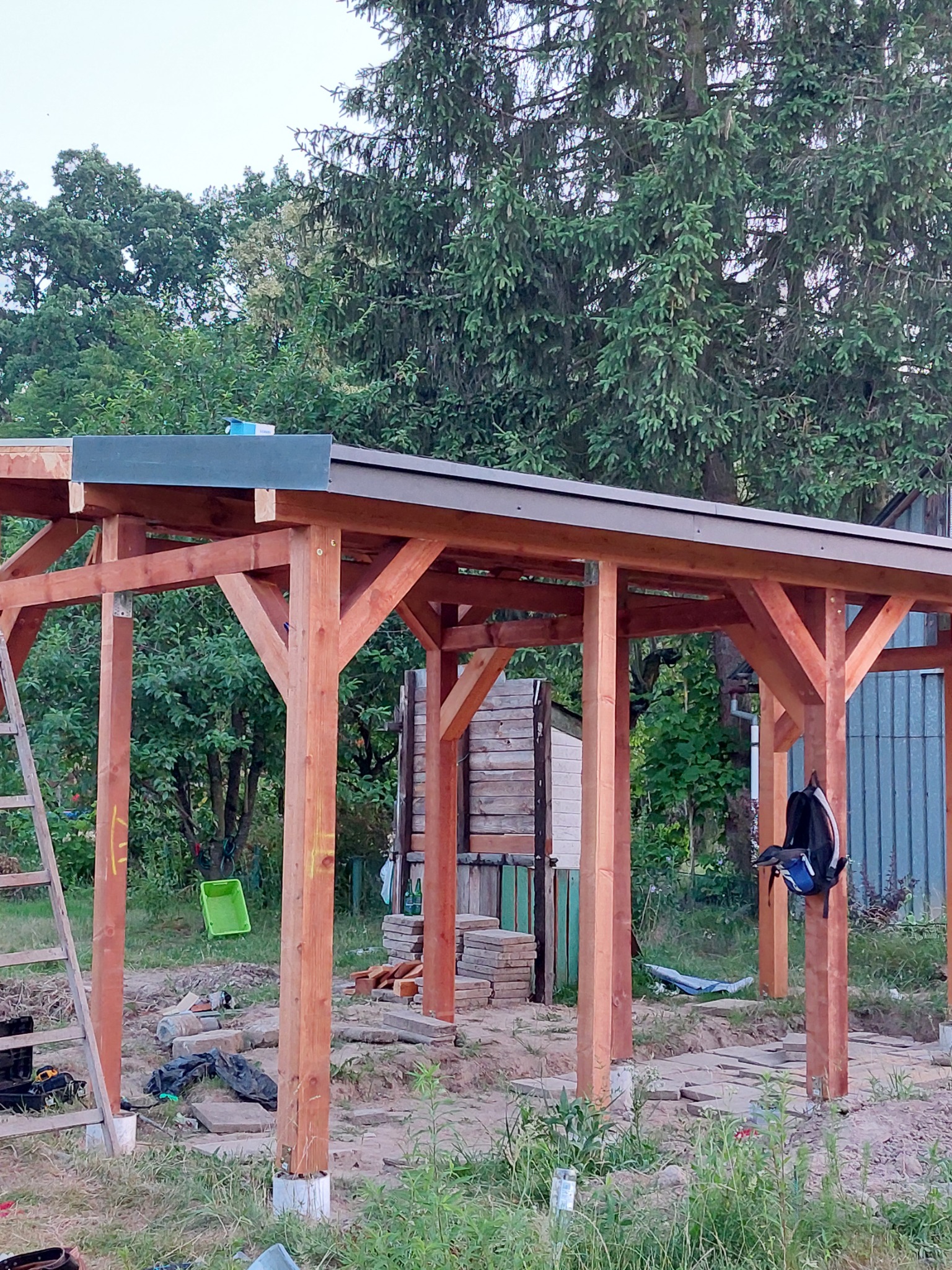 Konstrukcja drewnianej altany z częściowo ułożonym dachem z blachy, widoczne elementy mocujące i otoczenie placu budowy z narzędziami i materiałami budowlanymi.