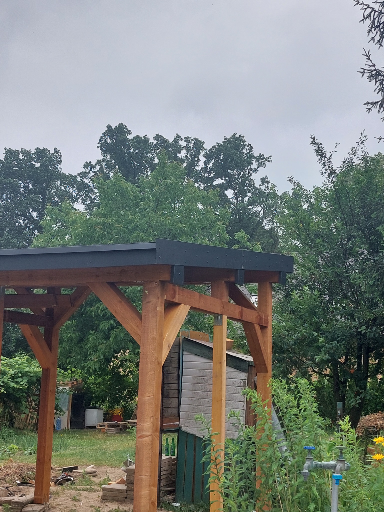 Drewniana konstrukcja z czarnym zadaszeniem w ogrodzie, widoczny fragment szopy i roślinność, pochmurne niebo w tle.