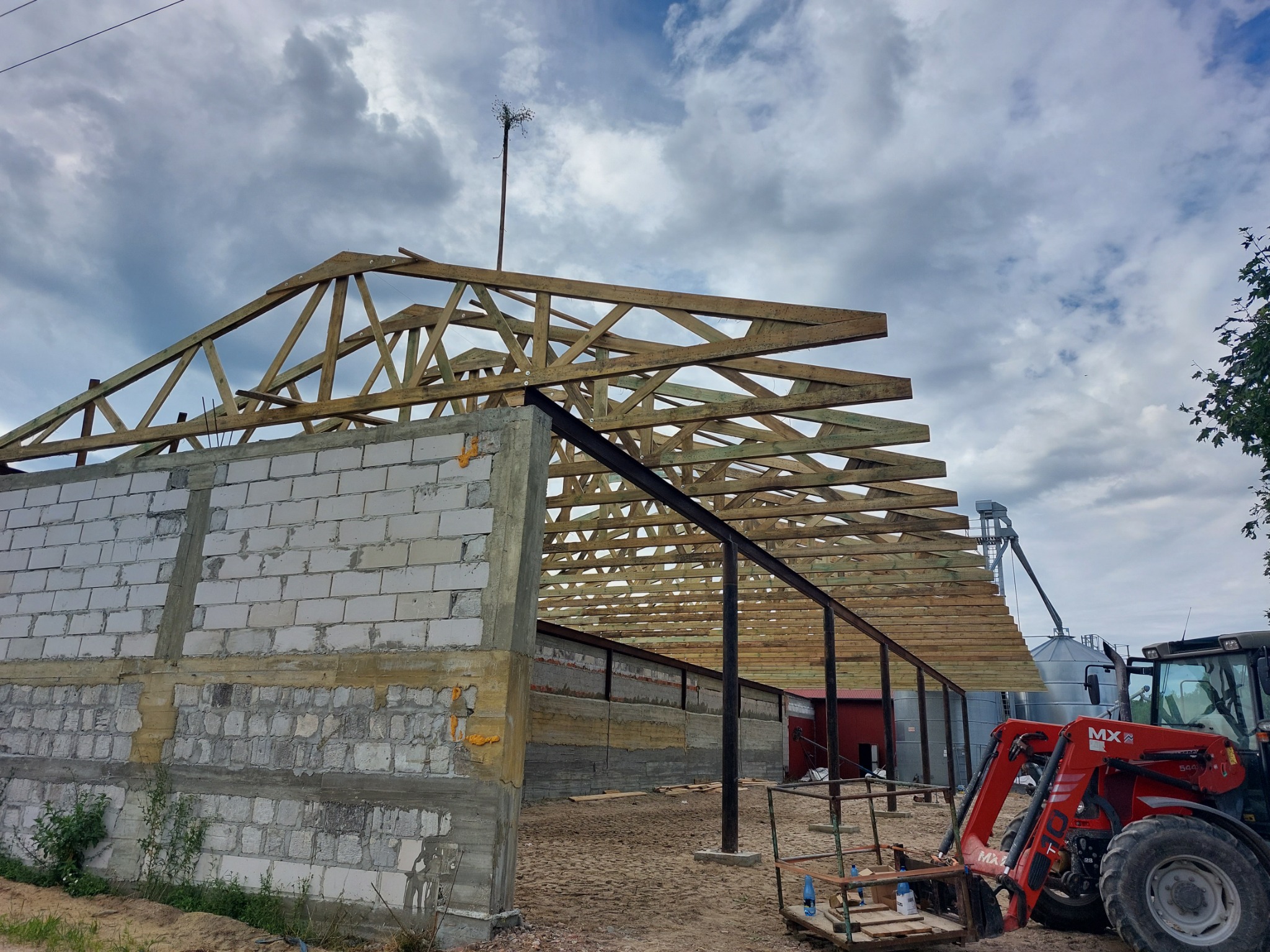 Konstrukcja drewniana dachu budynku gospodarczego w trakcie budowy, widoczny szkielet dachu i ściany z bloczków, na zewnątrz czerwony ciągnik rolniczy z podnośnikiem.