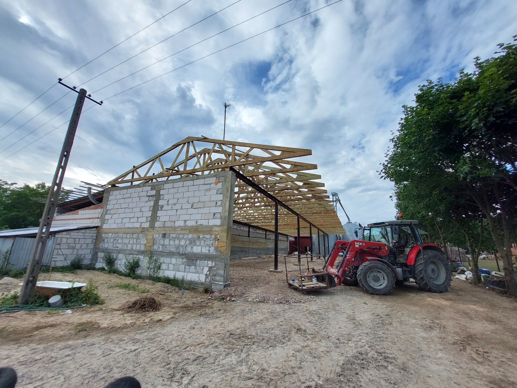 Budowa wiaty z drewnianą konstrukcją dachu, widoczny czerwony traktor z przodu i słup wysokiego napięcia po lewej stronie.