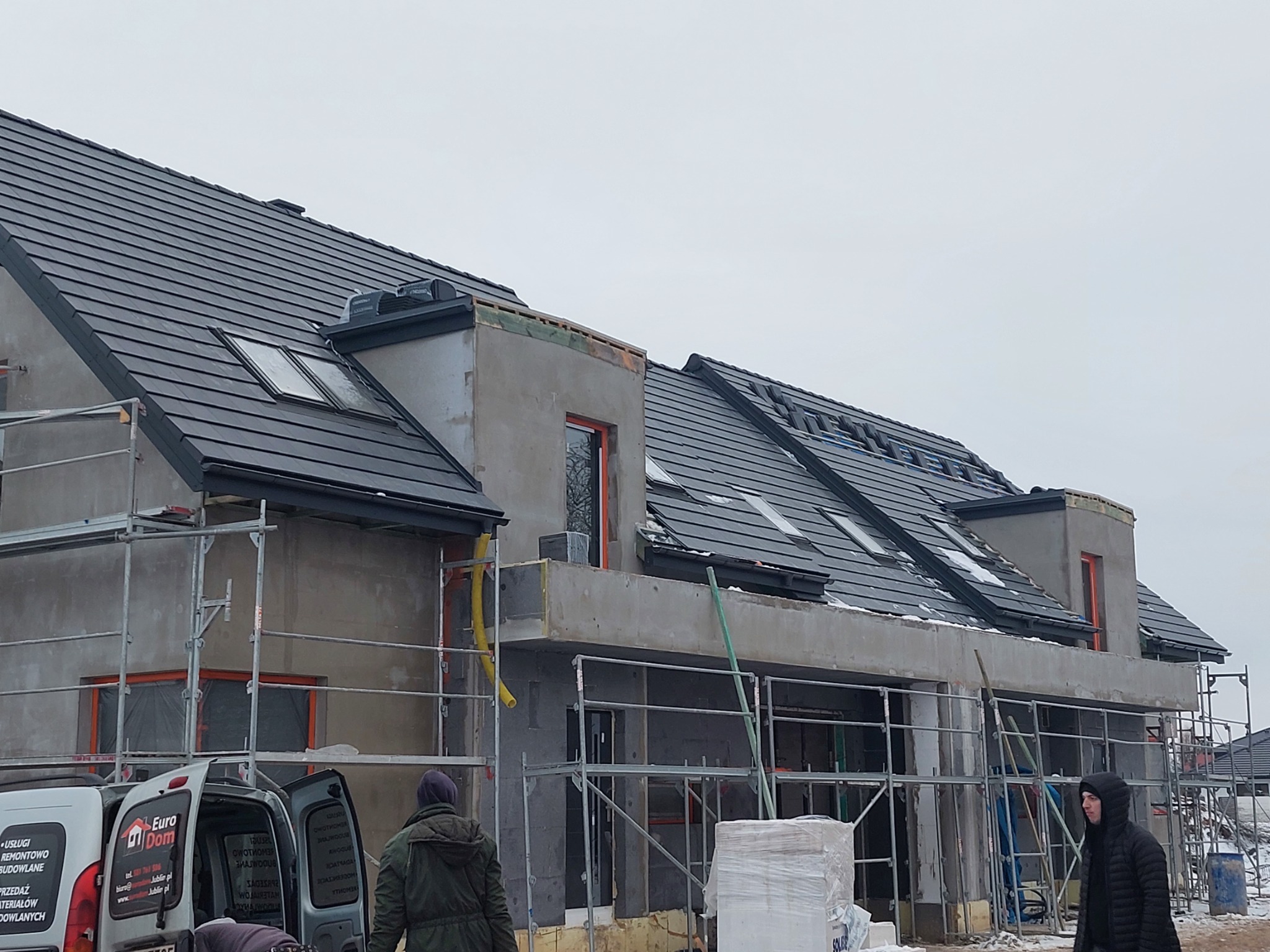 Nowy dom w trakcie budowy z ciemnoszarą dachówką, widoczne rusztowania, częściowo ośnieżony dach i teren, oraz samochód dostawczy z logo firmy budowlanej