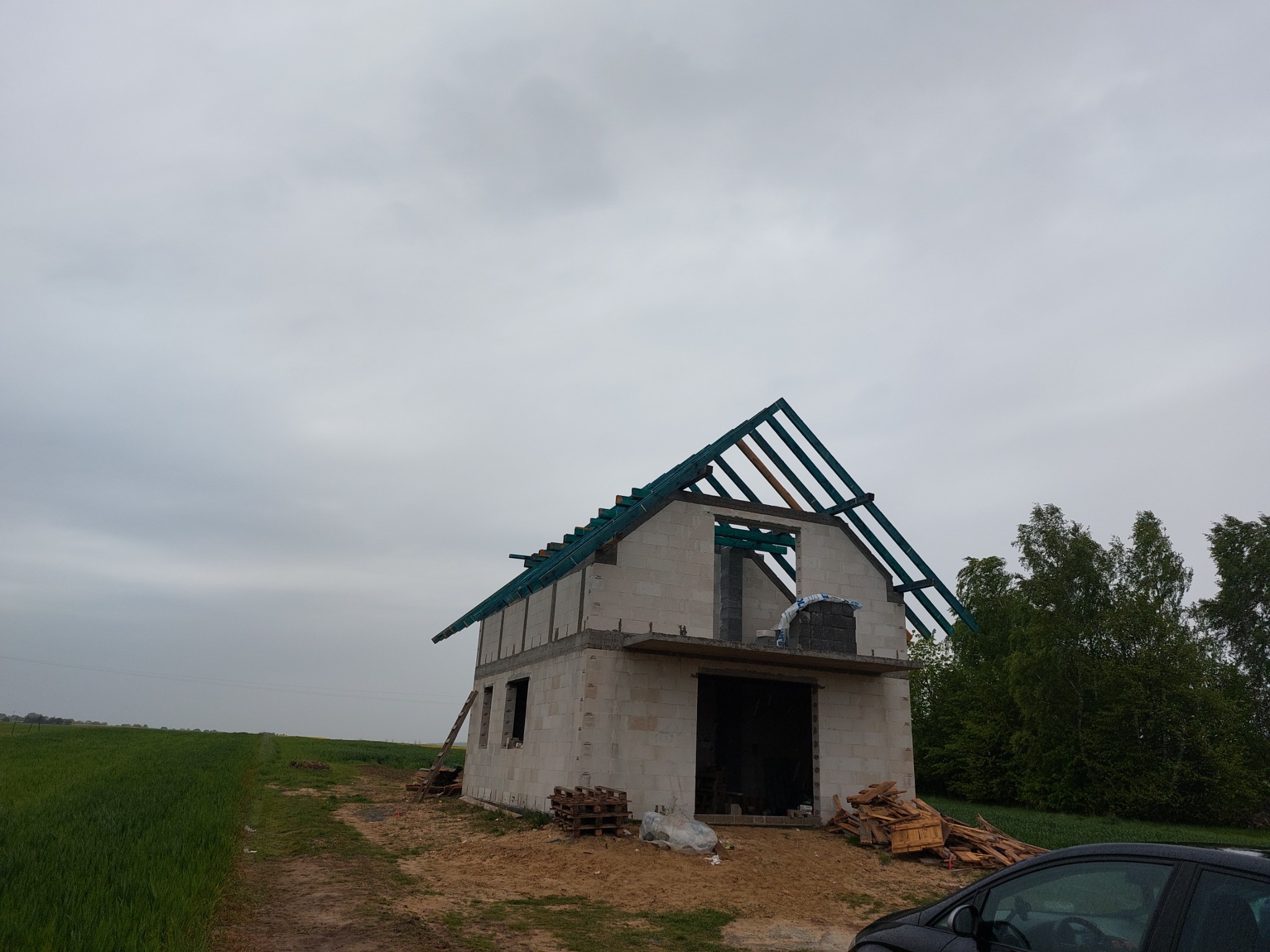 Surowy stan budynku jednorodzinnego z widoczną konstrukcją dachu w trakcie budowy, palety i drewno budowlane na placu budowy, pochmurne niebo w tle.