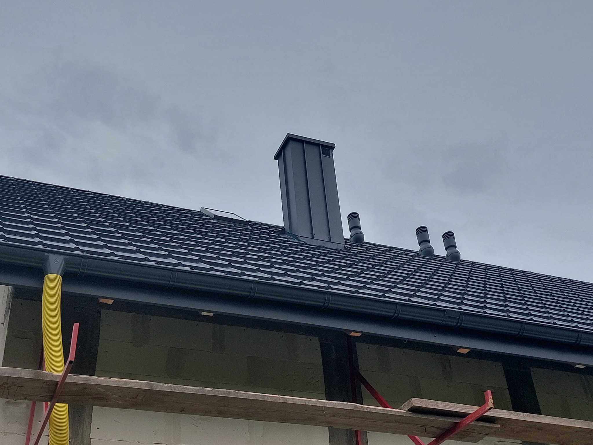 Czarny dach z metalowej dachówki z kominem i systemem wentylacji, widoczne żółte rury spustowe i fragment rusztowania.