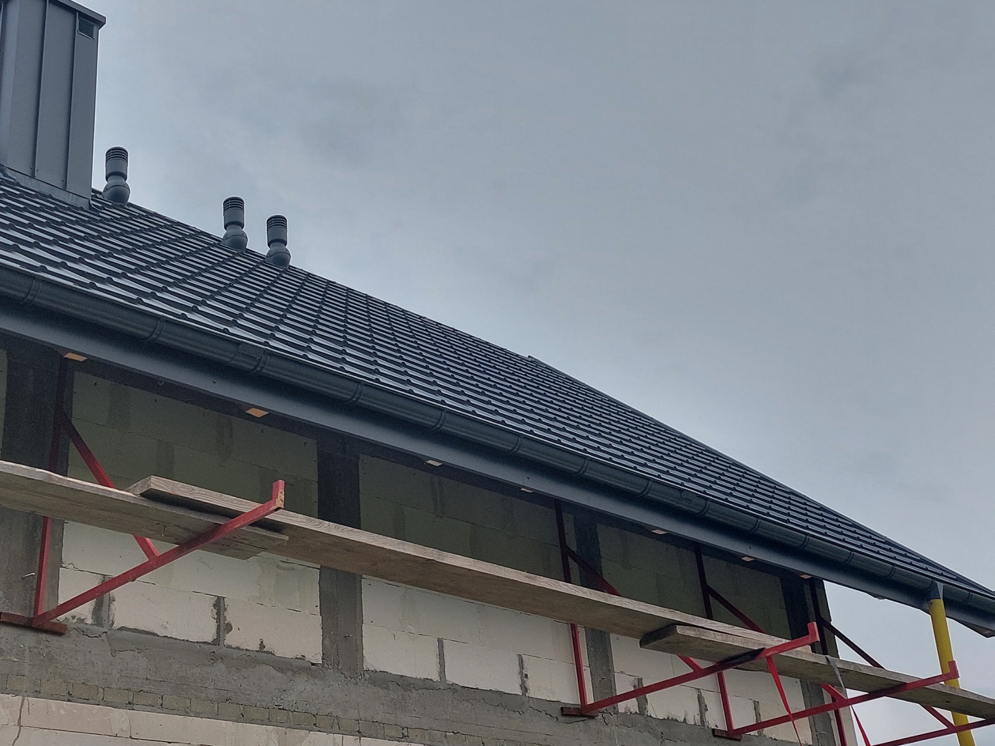 Nowy dach z ciemnej dachówki ceramicznej, widoczne kominki wentylacyjne i rynna, budynek w trakcie budowy z widocznymi rusztowaniami.