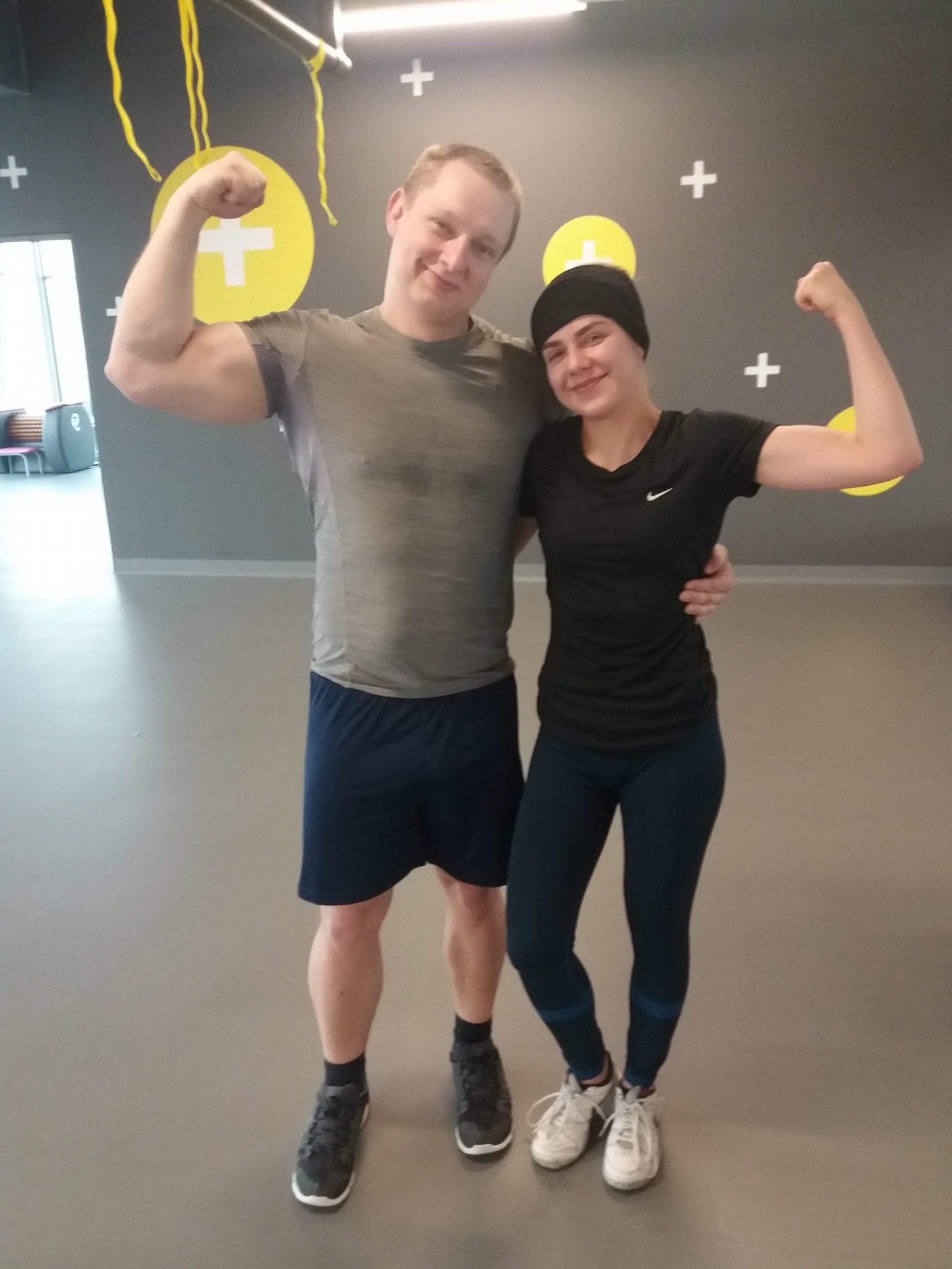 Uśmiechnięta para w sportowych strojach, prezentująca bicepsy na tle szarej ściany z żółtymi akcentami w siłowni.