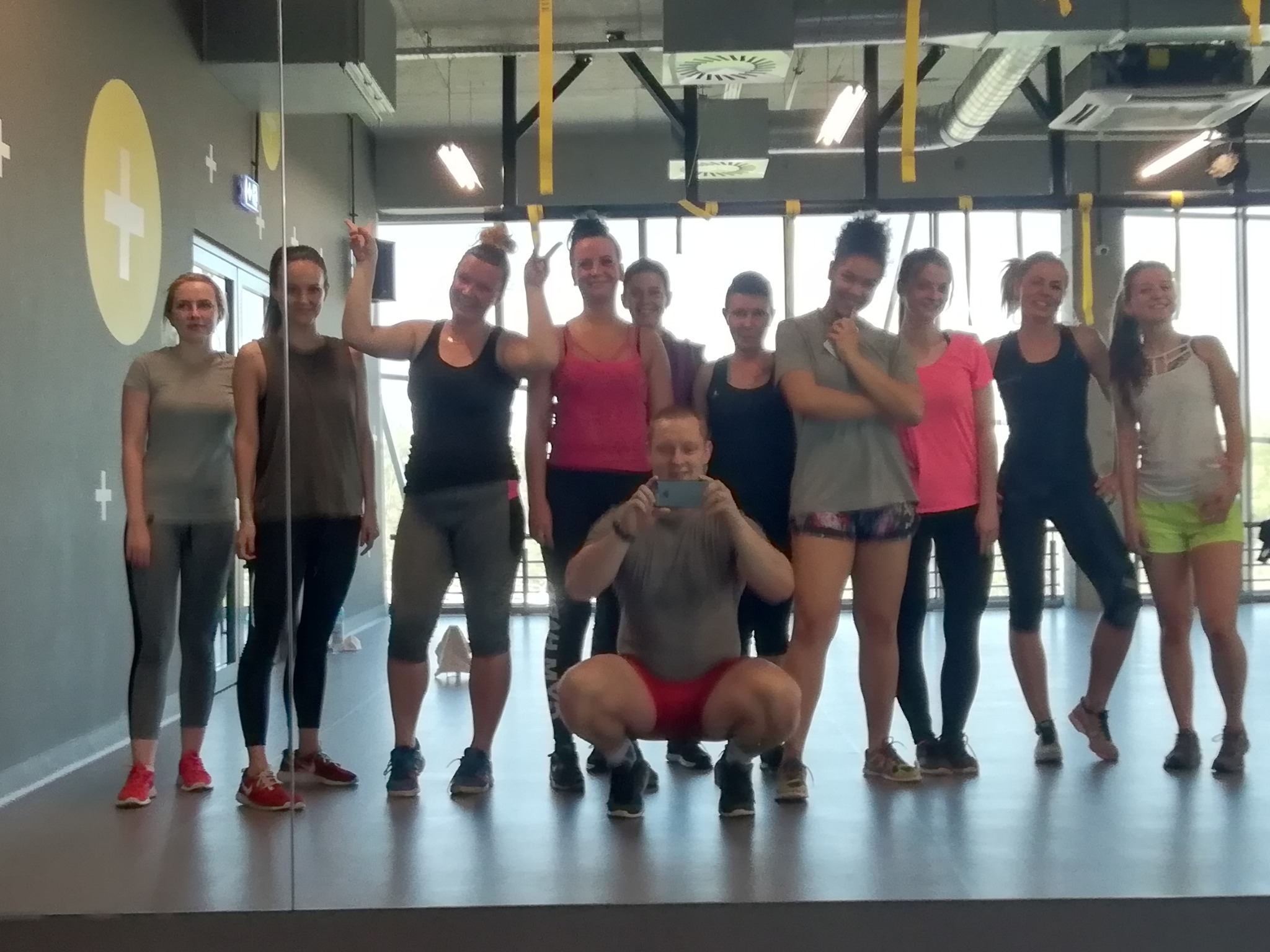 Grupa kobiet i mężczyzna w przysiadzie robiący zdjęcie telefonem, w tle sala fitness z żółtymi akcentami.