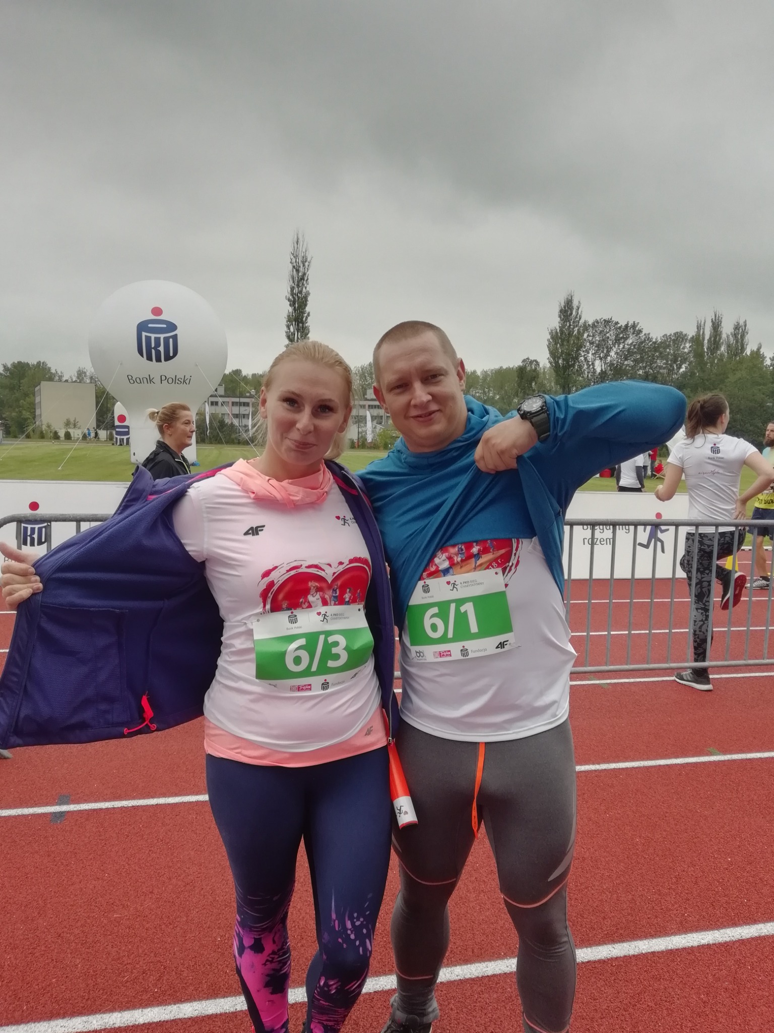 Dwoje biegaczy pozuje na bieżni po biegu, z numerami startowymi 6/1 i 6/3, w tle balon reklamowy Banku Polski.