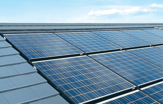 Panel słoneczny zamontowany na ciemnoszarym dachu z dachówki ceramicznej, widok z góry pod lekkim kątem na niebieskie niebo z delikatnymi chmurami.