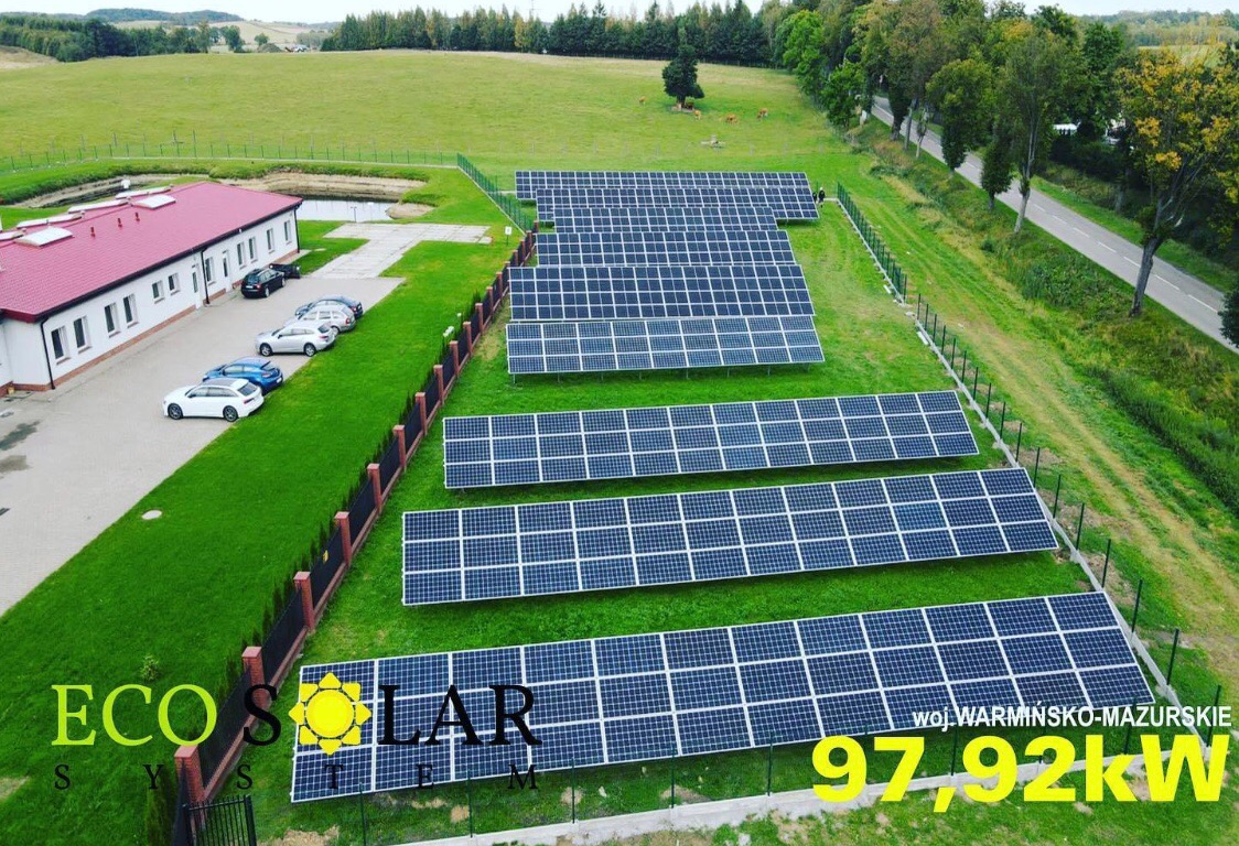 Widok z góry na instalację paneli słonecznych na trawiastym terenie, z budynkiem z czerwonym dachem i zaparkowanymi samochodami w tle, obok drogi i drzew.