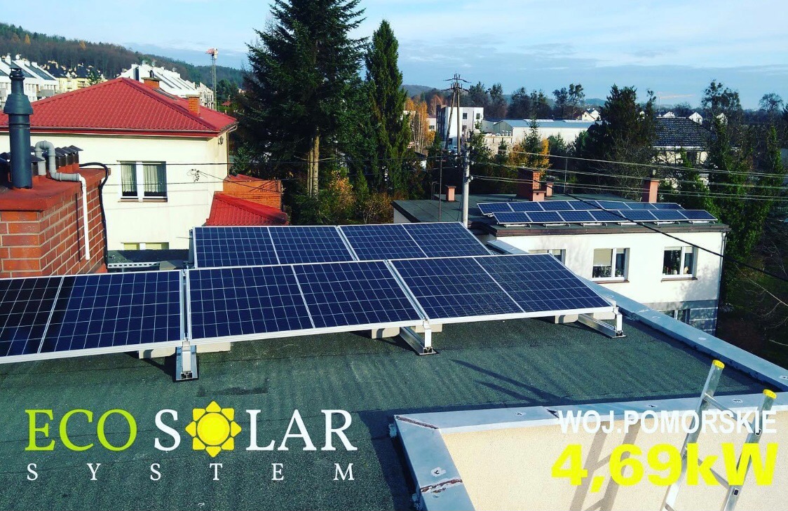 Instalacja paneli słonecznych na płaskim dachu budynku z widocznym logo firmy 'Eco Solar System' i informacją o mocy 4.69kW, w tle zabudowa mieszkalna i drzewa.