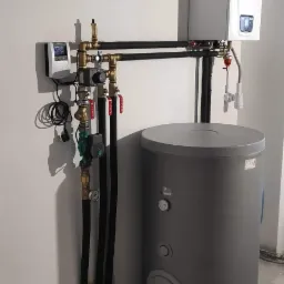 Zestawienie: biały piec gazowy kondensacyjny, szary zasobnik ciepłej wody, czarne rury instalacyjne, mosiężne zawory i sterownik na ścianie w surowym wnętrzu.