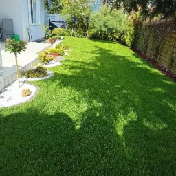 Zadbany trawnik przydomowy z równo przystrzyżoną trawą, ozdobnymi krzewami i drzewkami, wykończony białym kamieniem dekoracyjnym i drewnianym płotem z roślinnością.