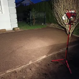 Przygotowany teren pod trawnik z rolki oświetlony halogenem, widoczne obrzeża i dwie rolki trawy przy budynku.