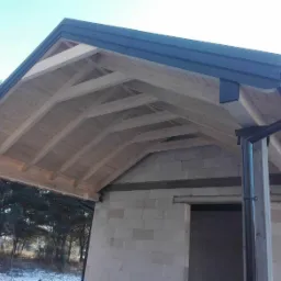 Surowa konstrukcja drewniana dachu z zamontowanymi rynnami i obróbkami blacharskimi, widoczna na tle częściowo wykończonego budynku z bloczków betonowych i zimowego krajobrazu.