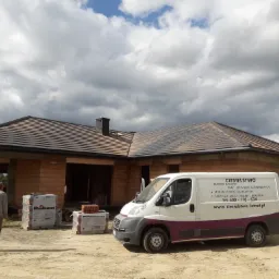 Dom w trakcie budowy z częściowo wykończonym brązowym dachem, obok zaparkowany biały bus z reklamą firmy ciesielskiej, na pierwszym planie stosy dachówek Roben, na ziemi piasek i pracownik budowlany.