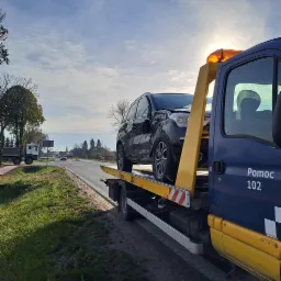 Uszkodzony czarny samochód osobowy na żółtej platformie pojazdu pomocy drogowej 'Pomoc 102', widok z boku na tle drogi i pochmurnego nieba.