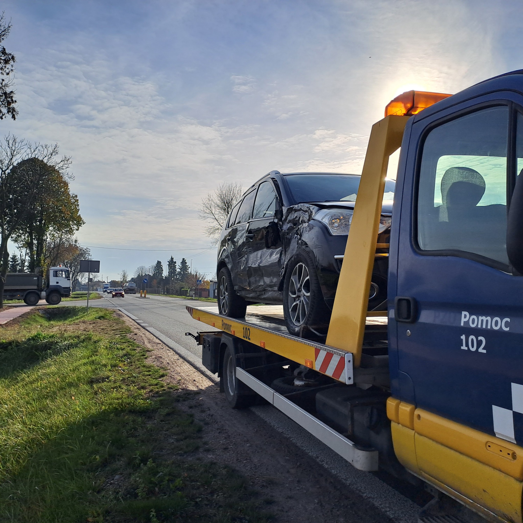 Uszkodzony czarny samochód osobowy na żółtej platformie pojazdu pomocy drogowej 'Pomoc 102', widok z boku na tle drogi i pochmurnego nieba.