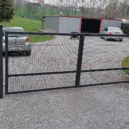 Otwarta, dwuskrzydłowa brama wjazdowa z metalowej siatki, pomalowana na ciemny kolor, widoczna na żwirowym podjeździe w tle z samochodami i budynkiem gospodarczym.