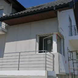 Nowoczesny dom w trakcie wykończenia z balkonami zabezpieczonymi prostymi, poziomymi barierkami ze stali nierdzewnej, widoczne rusztowanie przy elewacji.