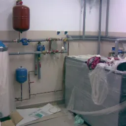 Kotłownia w trakcie prac instalacyjnych: zbiornik ciśnieniowy, bojler, piec owinięty folią ochronną i rury.