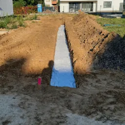 Wykop fundamentowy wyłożony białą geowłókniną, widoczny cień osoby, w tle dom jednorodzinny i błękitne niebo.