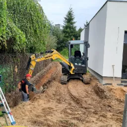 Żółta mini koparka Yanmar w trakcie prac ziemnych w Łukowie, operator w czerwonej czapce, obok niwelator laserowy, widoczny wykop pod fundament.